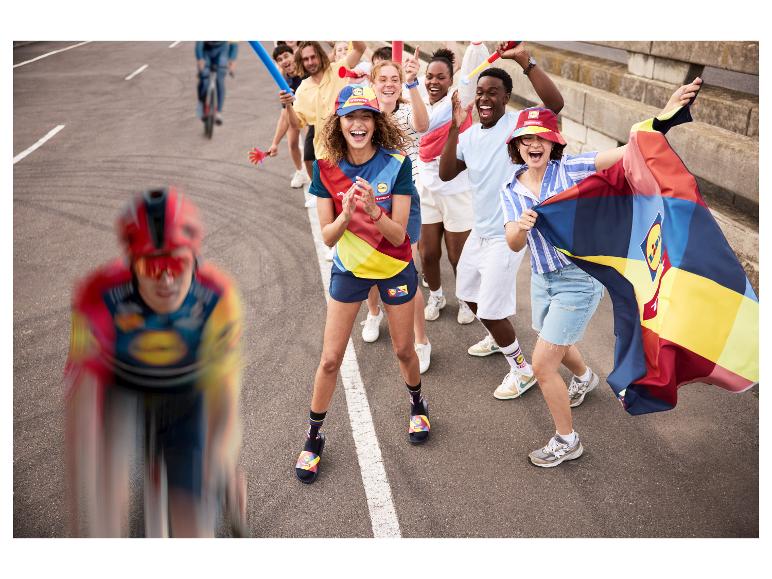 Radfahrer und jubelnde Fans in bunter Sportkleidung, Mützen und Socken mit einer Flagge.