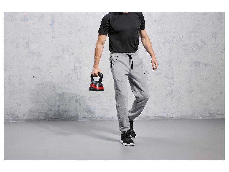 Mann in grauen Jogginghosen und schwarzen Sneakers, mit einer Kettlebell in der Hand.