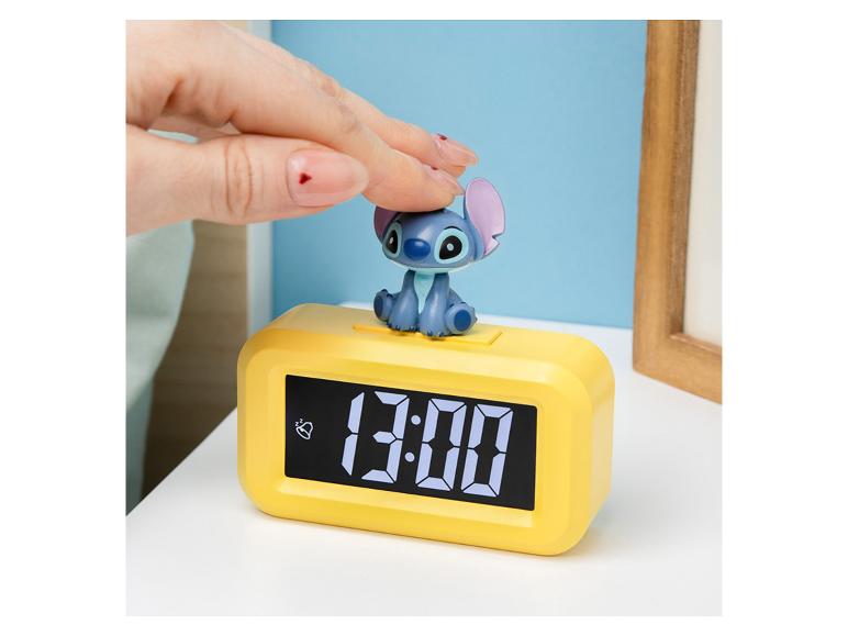 Eine Hand drückt auf eine Stitch-Figur auf einem gelben Digitalwecker, der „13:00“ anzeigt.