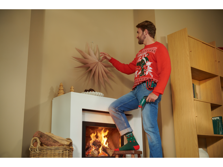 Mann im Weihnachtspullover dekoriert das Zimmer mit einem Akkuschrauber in der Hand.
