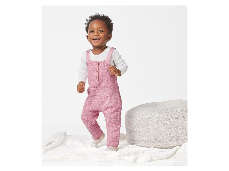 Ein Kleinkind in einem rosafarbenen Strick-Strampler spielt auf einer weißen Decke.