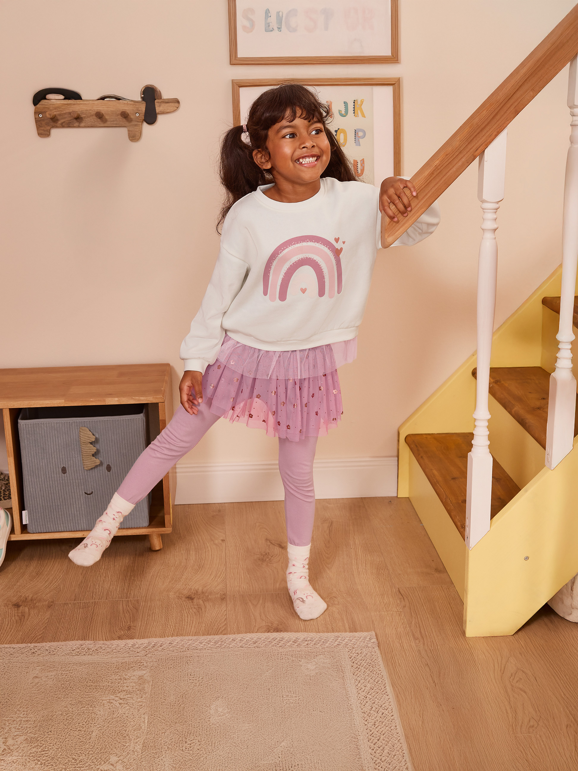Mädchen in rosa Tüllrock und Regenbogen-Sweatshirt, an einem Treppengeländer lehnend.