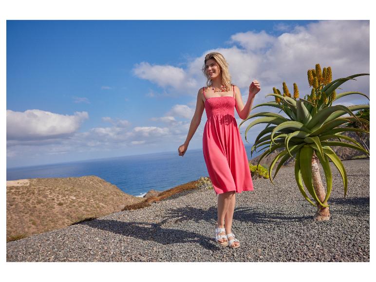 Frau in einem pinkfarbenen Sommerkleid und Sandalen, steht draußen am Meer mit einer Aloe-Pflanze.