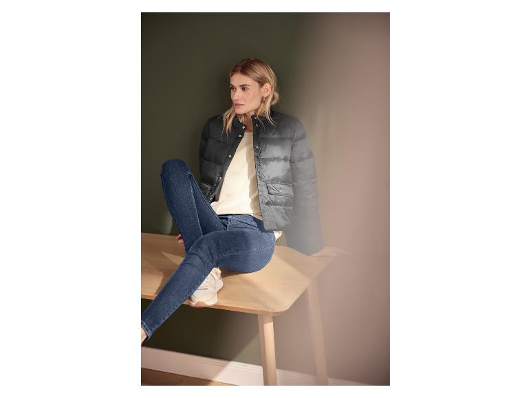 Frau in schwarzer Steppjacke, cremefarbenem T-Shirt und blauen Jeans, sitzt auf einer Holzbank.