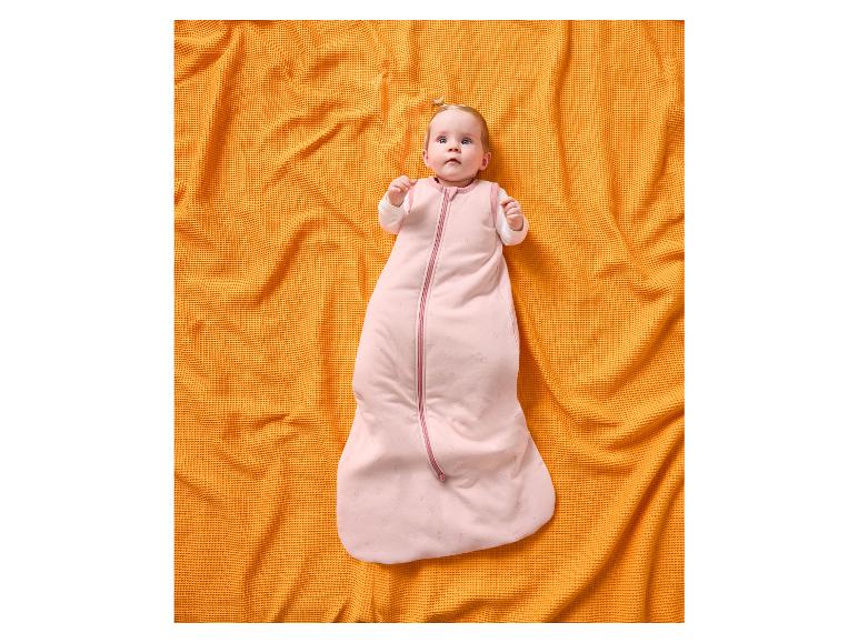Baby in einem rosa Schlafsack mit Sternen, liegend auf einer orangefarbenen Waffeldecke.