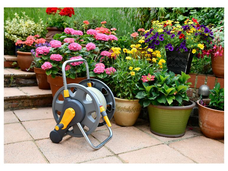 Grauer und gelber Gartenschlauchwagen auf einer sonnigen Terrasse mit bunten Topfpflanzen.