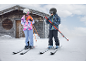 Zwei Kinder in Skianzügen, Helmen und auf Skiern, vor einer Holzhütte.