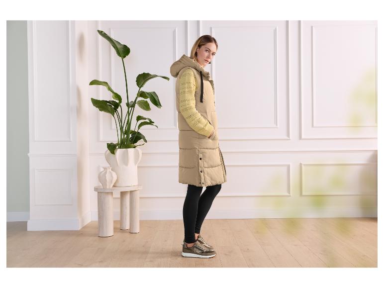 Frau in langer beiger Steppweste und gelbem Strickpullover, steht drinnen.