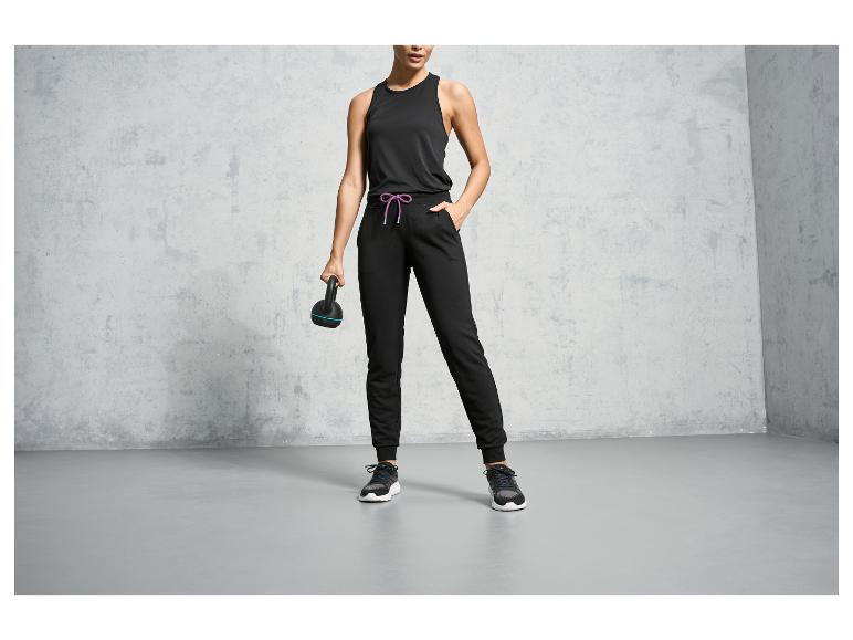 Eine Frau in schwarzen Jogginghosen, einem Tanktop und Sneakers hält eine Kettlebell.