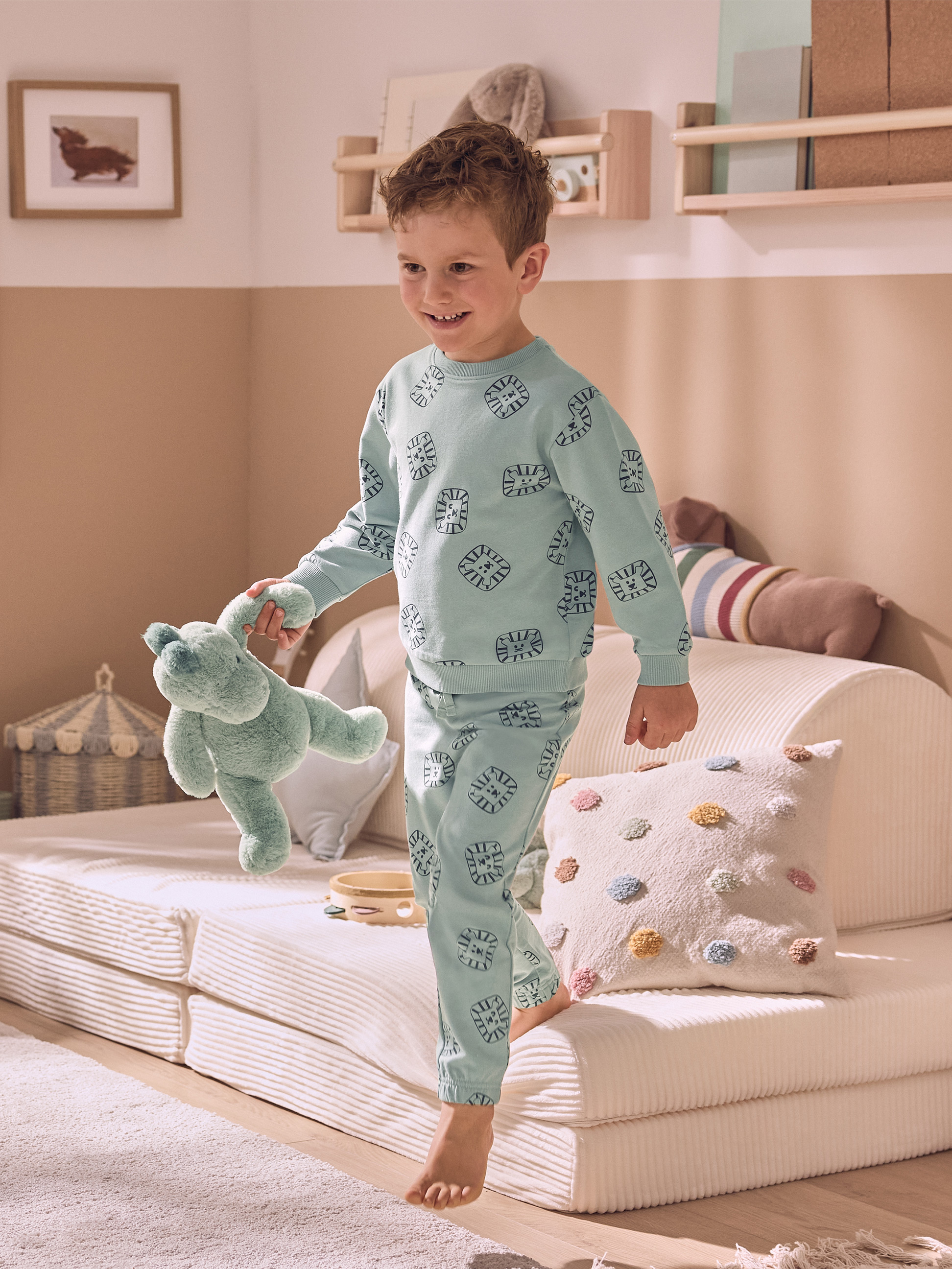 Ein lächelndes Kind in hellblauem Pyjama mit Löwenmotiven, das ein grünes Plüschtier hält.