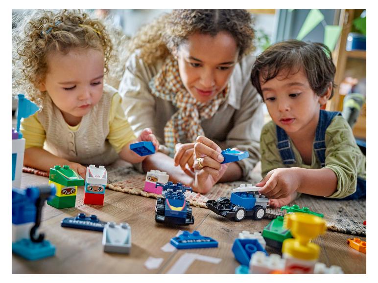 Erwachsener und zwei Kinder spielen mit bunten LEGO Duplo Steinen auf dem Boden.