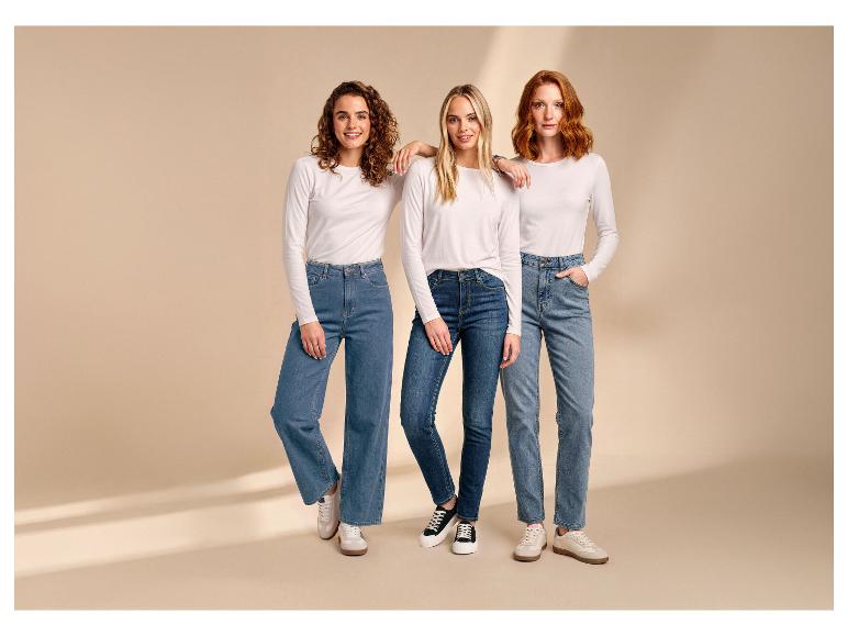 Drei Frauen tragen weiße Langarmshirts und Jeans, posieren vor beigem Hintergrund.