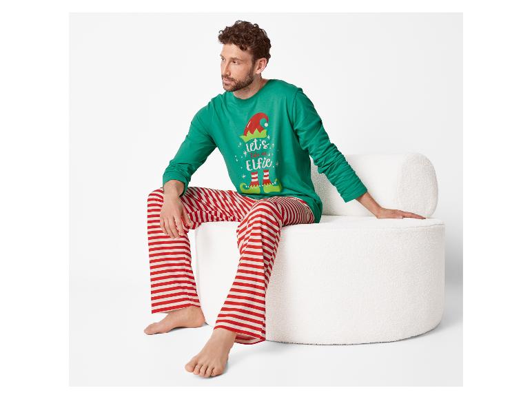 Mann sitzt auf weißem Stuhl in grünem Weihnachtspyjama mit „let's take an Elfie“-Aufschrift und rot-weiß gestreiften Hosen.