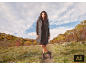 Frau in einer langen grauen Weste, einem braunen Kleid und Stiefeln in einer Herbstlandschaft.