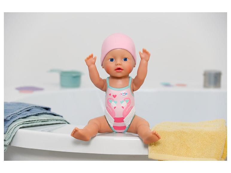 Babypuppe im rosa Badeanzug und Mütze, sitzt auf dem Badewannenrand mit Handtüchern.