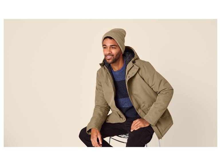 Mann in olivgrüner Jacke, blauem Pullover und Mütze, sitzt auf einem Hocker.