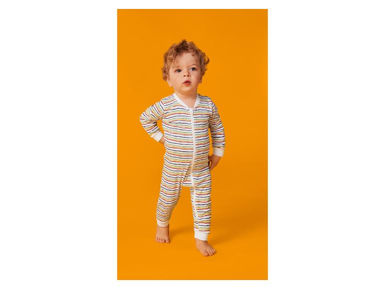 Lockiges Baby in gestreiftem Strampler steht vor orangefarbenem Hintergrund.