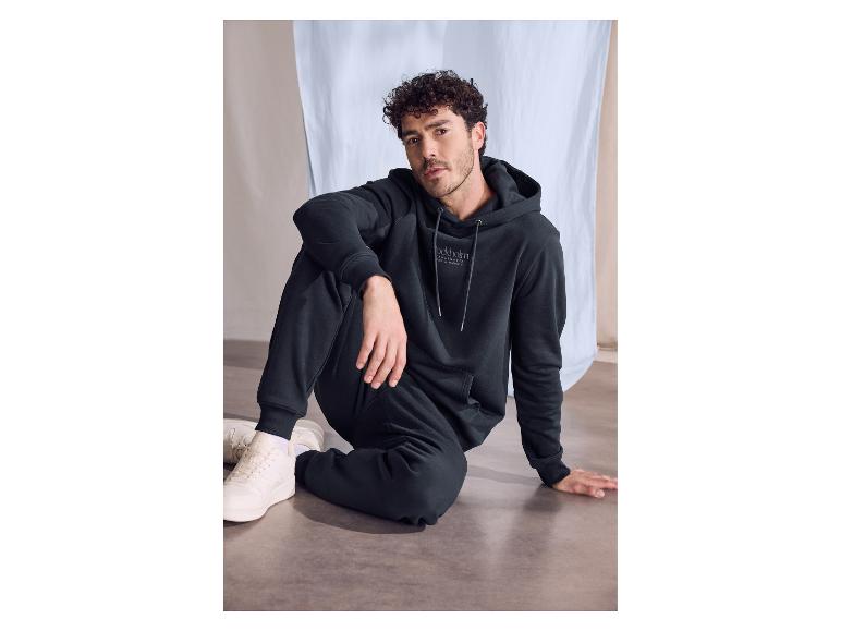 Mann mit dunklen Locken in schwarzem Kapuzenpullover und Jogginghose, auf dem Boden sitzend.