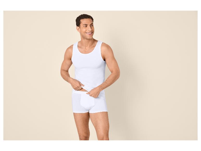 Mann in weißem Unterhemd und Boxershorts, lächelnd und zur Seite blickend.