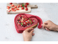 Herzförmiger Käsekuchen mit Beeren in einer roten Silikonform, mit Erdbeeren auf einem Schneidebrett.