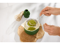 Eine Person siebt grünes Matcha-Pulver in eine Schale auf einem Holzuntersetzer.