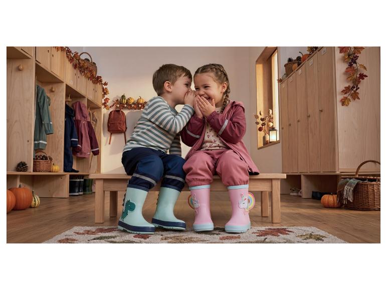 Zwei Kinder in Regenanzügen und Gummistiefeln flüstern Geheimnisse in einer herbstlichen Garderobe.