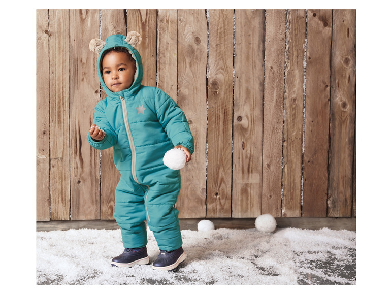 Ein Kind in einem grünen Winteroverall mit Öhrchen spielt mit Schneebällen.