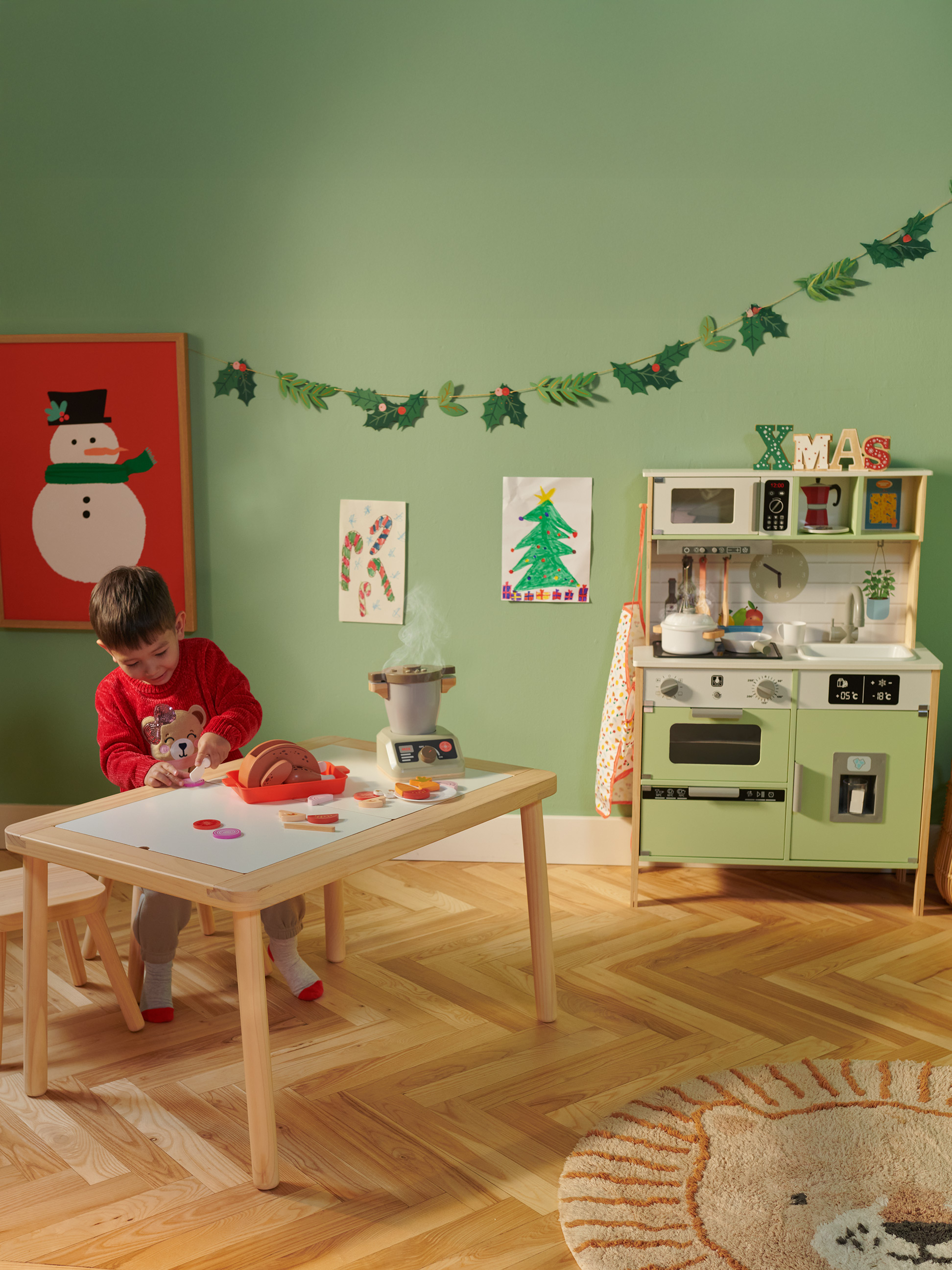 Ein Kind spielt mit einer Spielküche und einem Spielzeug-Esstisch in einem weihnachtlich dekorierten Raum.