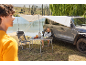 Frau sitzt an Klapptisch und Stuhl unter Markise am Auto, am See.