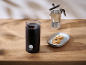 Schwarze Kaffeemühle mit Bohnen, Mokkakanne und Kekse auf einem Holztisch.
