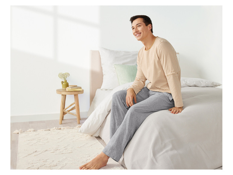 Mann in gestreiftem Pyjama und grauer Hose, sitzend auf einem Bett.