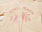 Ein beiges T-Shirt mit dem Aufdruck 'Sunset Beach' und Palmen.