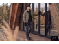 Mann in khakifarbener Steppjacke, schwarzem Pullover, Jeans und Mütze, auf Holzterrasse.