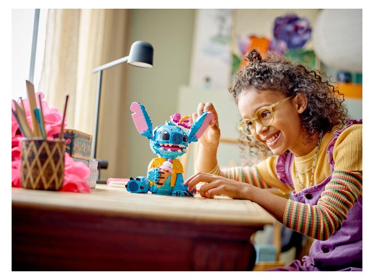 Ein lächelndes Mädchen spielt mit einer großen Stitch-Figur aus LEGO-Steinen auf einem Tisch.