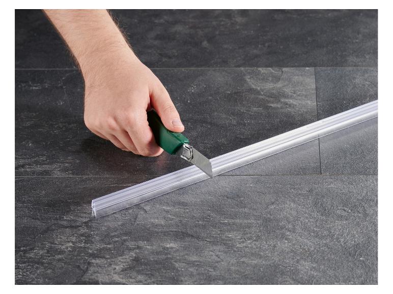Hand schneidet eine transparente Duschdichtung mit einem Cuttermesser auf Fliesenboden.