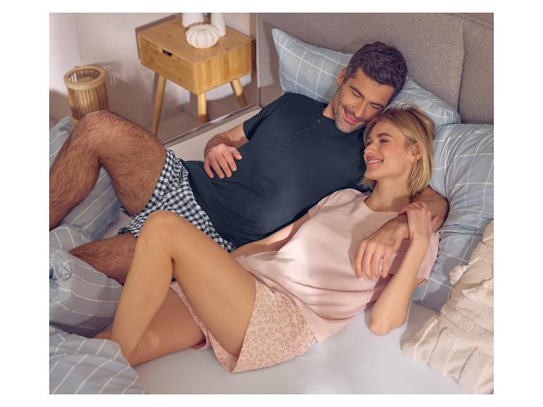 Lächelndes Paar im Pyjama im Bett, Mann in dunklem Oberteil und karierten Shorts, Frau in rosa Oberteil und floralen Shorts.