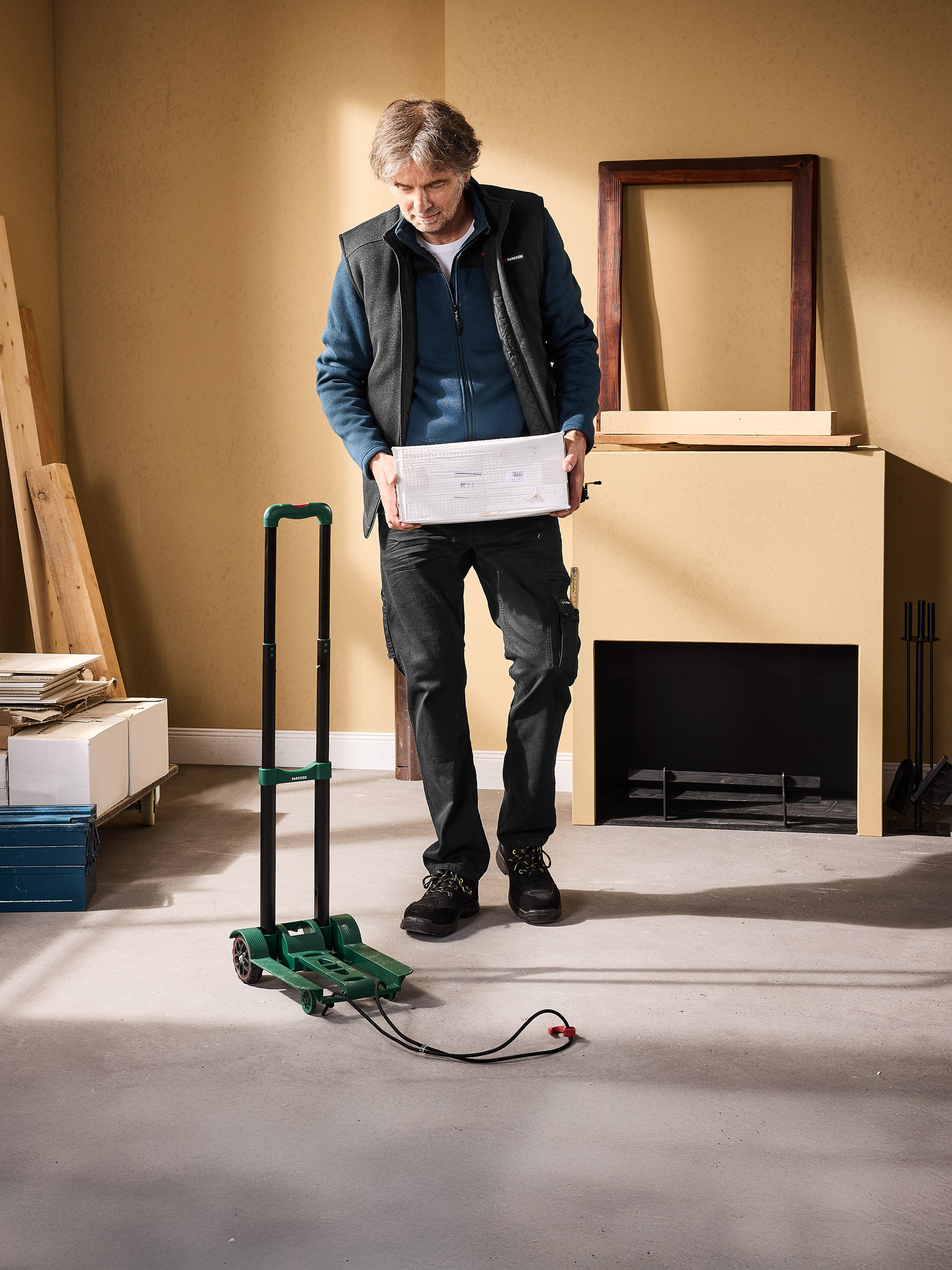 Mann hält eine weiße Kiste, neben einem grünen Rollwagen und einem Kamin.