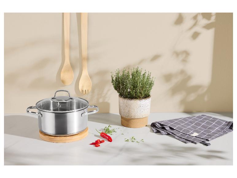 Edelstahltopf mit Glasdeckel auf Holzuntersetzer, mit Küchenutensilien und Kräutern.