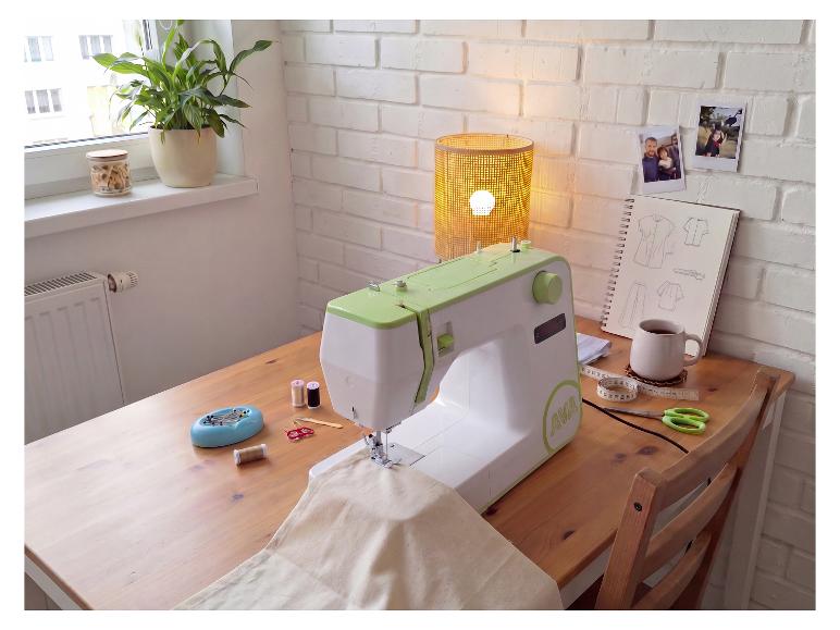 Nähmaschine auf einem Holztisch mit Nähzubehör, einer Pflanze und einer Lampe.