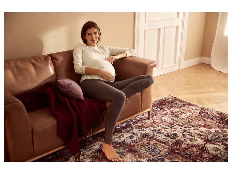 Eine schwangere Frau in einem weißen Langarmshirt und grauen Leggings sitzt auf einem braunen Ledersofa.