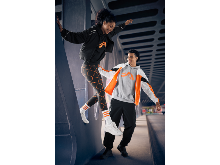 Zwei Personen in Sportkleidung, schwarzen und orangefarbenen Leggings, Hoodies und Jacken, lächelnd im Freien.