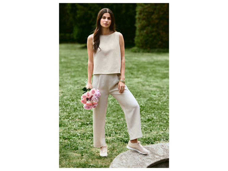 Eine Frau trägt ein beiges Oberteil und eine Hose mit weißen Sneakers in einem grünen Garten.