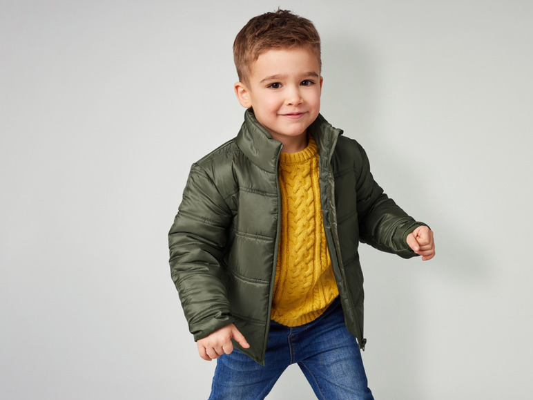 Ein Junge in einer grünen Jacke, einem gelben Pullover und blauen Jeans.