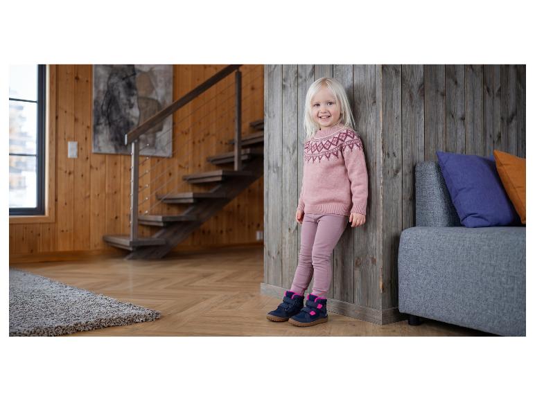 Lächelndes Mädchen in rosa Pullover und Hose, mit Stiefeln, an eine Holzwand gelehnt.