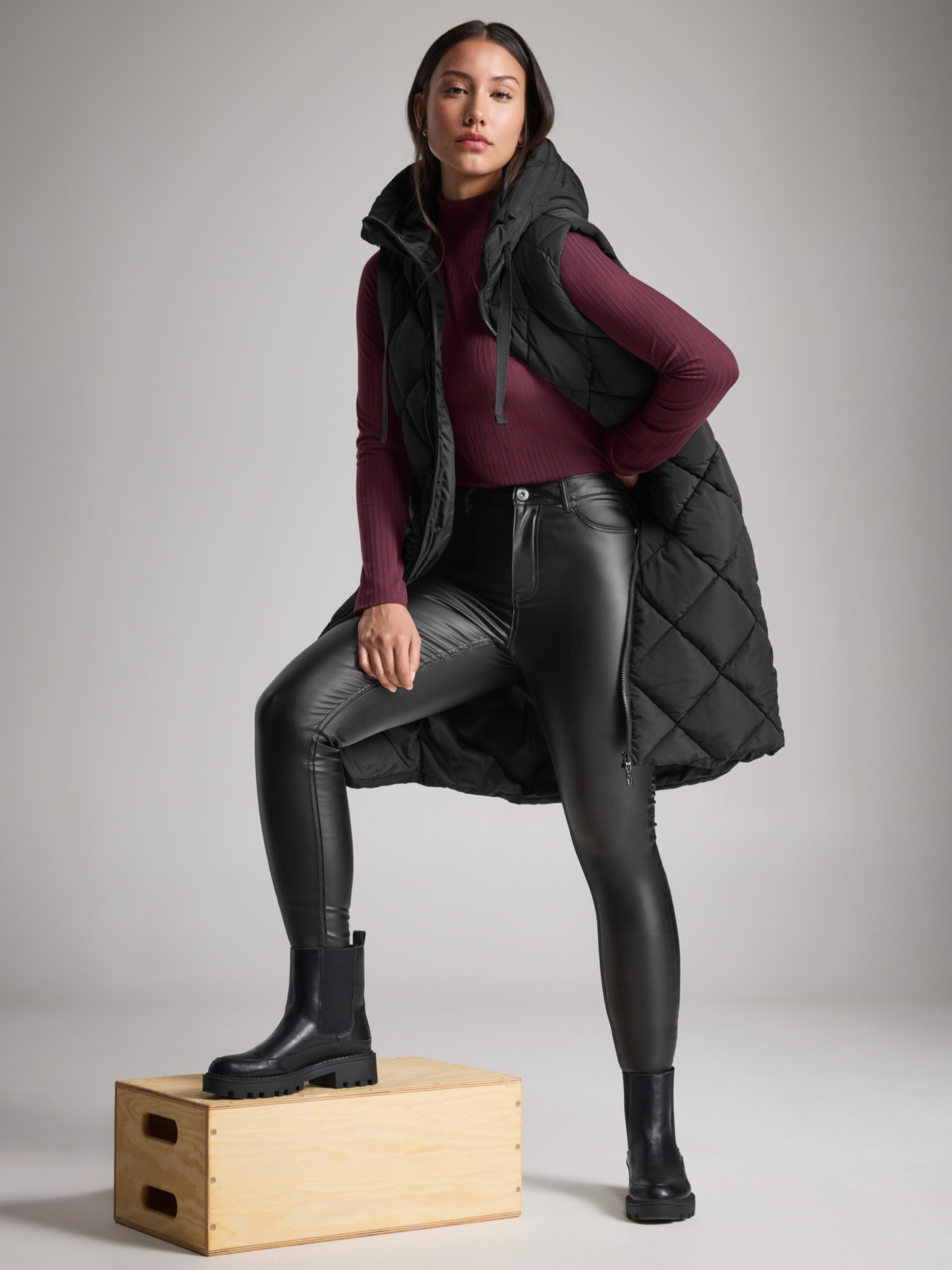 Frau in schwarzer Steppweste, Lederjeans und Stiefeln, mit einem Fuß auf einer Kiste.