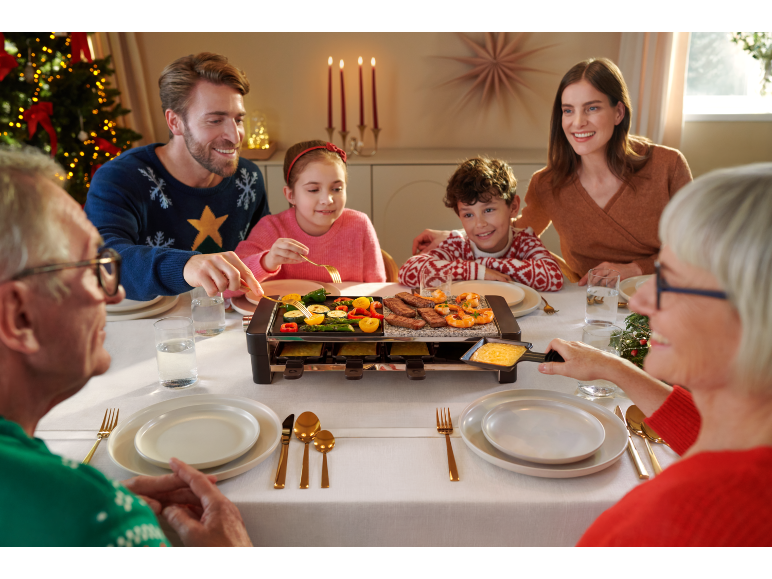 Familie am Weihnachtstisch versammelt, die auf einem Raclette-Tischgrill kocht.