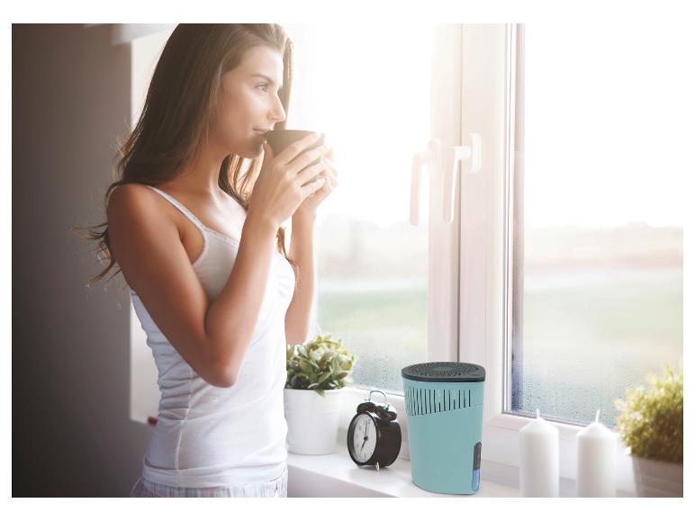Eine Frau trinkt Kaffee an einem beschlagenen Fenster, im Vordergrund ein Luftentfeuchter.