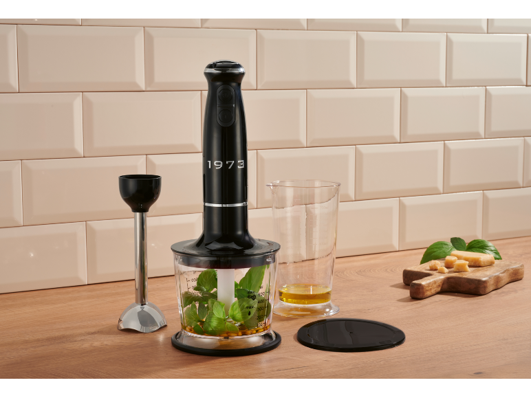 Schwarzer Stabmixer mit Zubehör, Kräutern und Käse auf einer Holztheke.