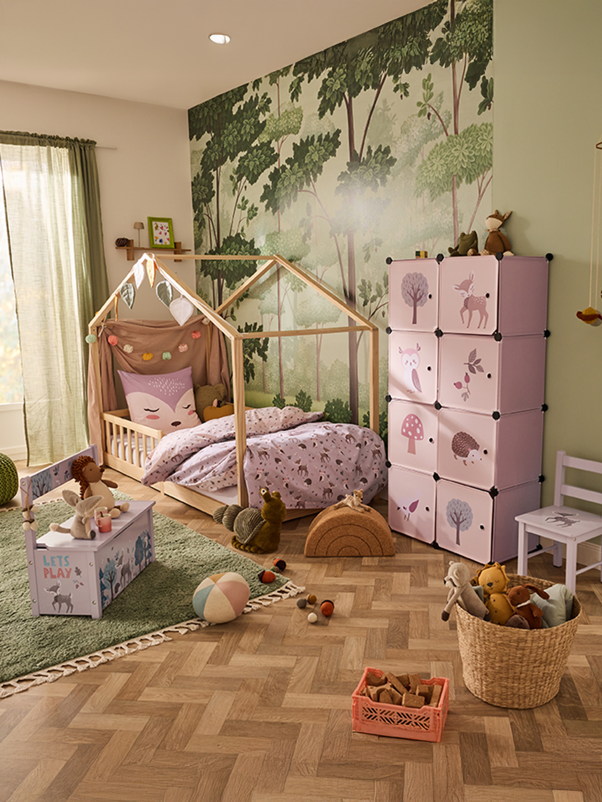 Kinderzimmer mit Hausbett, rosa Aufbewahrungswürfeln und Waldtapete.
