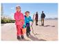 Kinder in Fleecejacken und Outdoorhosen spielen am Strand, Eltern im Hintergrund.